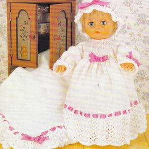 May include: A doll wearing a white knitted dress, bonnet, and blanket, all adorned with pink ribbon accents. The doll has blue eyes and blonde hair. A small wooden cabinet is in the background.