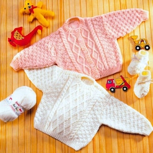 May include: A light pink and white knitted baby sweater with cable knit detailing. The sweaters are displayed with a yellow teddy bear, a red toy, a ball of yarn, and toy vehicles. White baby booties are also included.