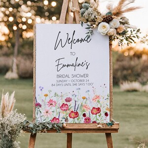 以下が含まれることがあります： エマラインのブライダルシャワーのウェルカムサインを表示する木製のイーゼル。サインには花のイラストと「Welcome to Emmaline's Bridal Shower Sunday - October 24 84 Days Until She Says I Do」というテキストが書かれています。シーンは屋外です。