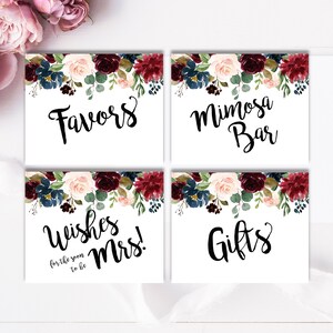 May include: Four white signs with floral borders in shades of burgundy, pink, and blue. The signs read "Favors", "Mimosa Bar", "Wishes for the soon to be Mrs.", and "Gifts".