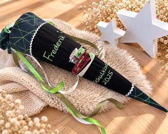 May include: A black cone-shaped school gift bag featuring a green geometric pattern at the top and bottom, the name Frederik, a red tractor graphic with the text CLAAS, and the text Schulkind 2025 in green, finished with green and white ribbons.