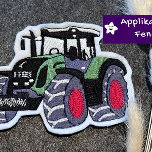 Könnte beinhalten: Gestickter Aufnäher eines grünen, schwarzen und blauen Traktors mit roten Radmitten. Der Aufnäher hat einen weißen Rand und enthält den Text "Applikation Fendt" in einer lila Box mit einem Stern.