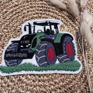 Könnte beinhalten: Gestickter Aufnäher mit einem grünen und schwarzen Traktor mit großen blauen Reifen und roten Naben. Der Traktor steht auf einer grünen Grasbasis und einem weißen Rand. Für Kleidung oder Accessoires.