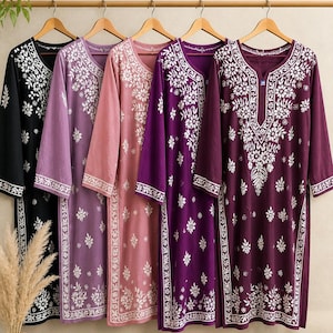 May include: Five long-sleeved tunics hanging on wooden hangers, each featuring a white floral embroidery pattern. The tunics come in black, light purple, dusty pink, bright purple, and deep plum, all with a notched neckline and side slits.