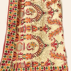 May include: Cream-colored textile with intricate embroidery in red, orange, green, and yellow. The design features floral and geometric patterns, with a red border. The fabric is draped over a wooden hanger.