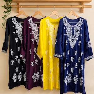 May include: Four long-sleeved tunics in black, purple, yellow, and navy blue hang on wooden hangers. Each tunic features intricate white floral embroidery around the neckline and sleeves. The tunics are displayed against a neutral background.