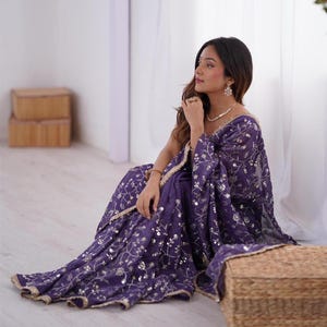 May include: A woman wearing a purple sari with silver floral embroidery and a gold border. The sari is draped around her, and she is seated on the floor. The sari's fabric has a shimmering effect, and the overall look is elegant and traditional.