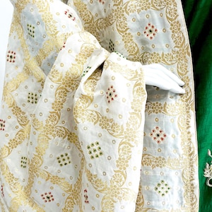 May include: A white shawl with a gold floral pattern and small, colorful embellishments. The shawl is draped over a green garment.