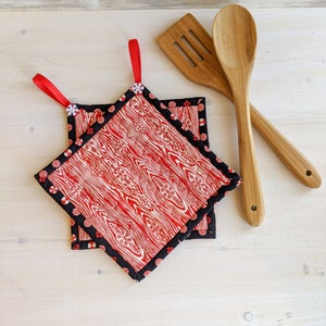 May include: Two red and white patterned pot holders with black trim and red fabric loops. The pot holders are stacked on top of each other. Two wooden spoons are lying on a white surface.