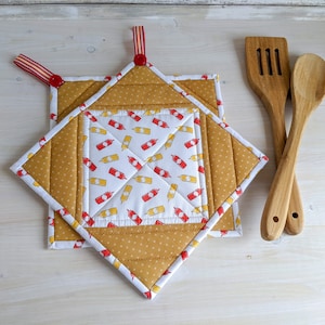 May include: Two quilted pot holders made from mustard yellow and white fabric with a pattern of red and yellow ketchup and mustard bottles. The pot holders have red ribbon loops for hanging.