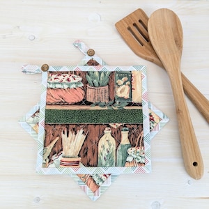 May include: Two square fabric pot holders featuring a kitchen shelf print with jars, plants, and utensils, bordered by a light blue and red geometric pattern. Each pot holder has a small wooden button at the top corner, paired with two wooden kitchen utensils.