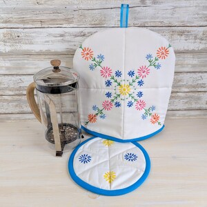 May include: A glass French press with a wooden handle alongside a white quilted tea cosy and a matching round trivet. Both fabric items feature embroidered floral patterns in orange, pink, yellow, and blue, finished with bright blue trim.