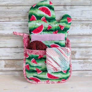 May include: A watermelon-themed kitchen set. It includes a green oven mitt with red watermelon slices, a pink pocket, a striped towel, a knitted scrubby, and a small square washcloth. Designed for kitchen use.