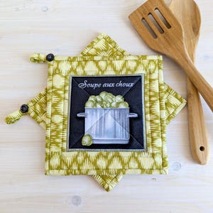 May include: A quilted pot holder with a green and white geometric pattern. The centre features a black square with a pot of cabbage and the text "Soupe aux choux". Two wooden cooking utensils are next to the pot holder.
