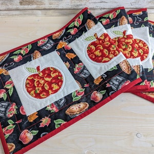 May include: Set of four black and white quilted table runners with red trim. Each runner features a white square with an applique of a red apple with green leaves. The background fabric has a pattern of apples, pies, and baskets.