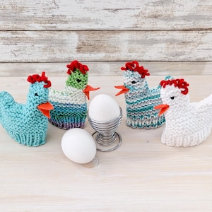 May include: Four knitted chicken egg cosies in solid light blue, variegated green and white, variegated blue and white, and solid white. Each features a red felt comb, orange felt beak, and black bead eyes. A silver wire egg cup holds one white egg.