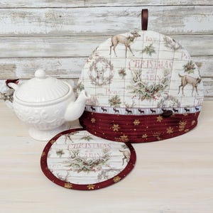 May include: A white ceramic teapot, a fabric tea cosy, and a matching round coaster. The tea cosy and coaster feature a cream background with deer illustrations, greenery, and the text Christmas at the Cabin, finished with a dark red border.