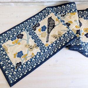 May include: A quilted table runner and two matching placemats. The runner and placemats feature a cream-coloured centre with a bird and floral design in blue, yellow, and black. The edges are bordered with a dark blue leaf pattern.