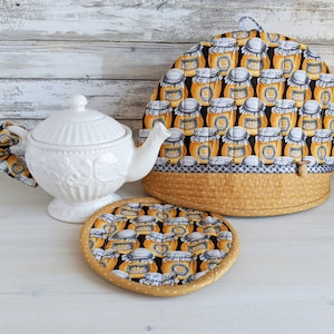 May include: A white ceramic teapot with a matching fabric tea cosy and a circular pot holder. The fabric features a repeating pattern of orange jars on a black background, with a mustard yellow polka dot border and a small wooden toggle detail.