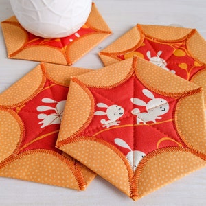 May include: Four orange and red fabric coasters with a white bunny pattern. The coasters have a scalloped edge and are quilted.