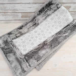 May include: A rectangular table runner featuring a black and white toile de jouy landscape print on the sides and a central panel with a white and grey snowflake pattern. The fabric has a quilted texture with stitched borders and edges.
