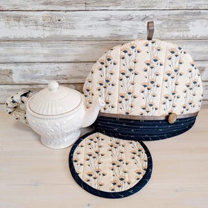 May include: A white ceramic teapot, a quilted tea cosy, and a matching round trivet. The tea cosy and trivet have a cream background with a floral pattern in blue and tan. The cosy has a navy blue base and a wooden button. The teapot is white.