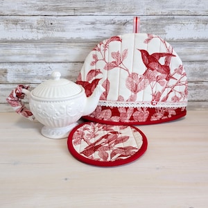 May include: A white ceramic teapot with a floral design, a matching tea cosy, and a coaster. The tea cosy and coaster feature a red bird and leaf pattern on a white background. The cosy has a red trim.