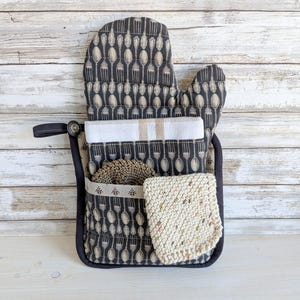 May include: A black oven glove with a utensil pattern, with a pocket containing a white and beige striped tea towel, a brown scrubby, and a cream-coloured knitted washcloth. A black loop and button detail are on the side.