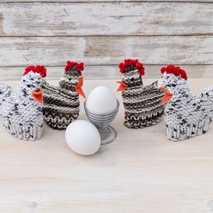 May include: Four knitted chicken-shaped egg warmers featuring speckled white and brown yarn, red felt combs, and orange felt beaks. Two are white with black speckles, and two are brown and white striped. A metal egg cup holds one white egg.