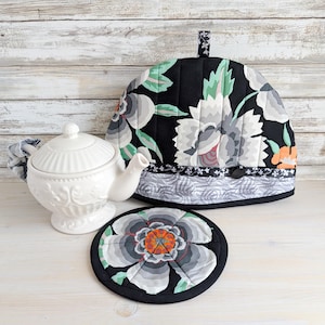 May include: A white ceramic teapot with a floral design sits beside a black tea cosy and a matching coaster. The tea cosy and coaster feature a black background with large grey, white, and orange floral patterns. The set is on a white wooden surface.