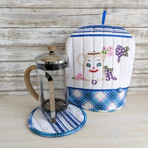 May include: A glass French press with a wooden handle sits on a round, white and blue striped quilted coaster next to a matching quilted tea cosy. The tea cosy features a blue plaid base, a blue hanging loop, and embroidered grape and teapot motifs.