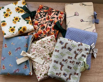 May include: A collection of fabric book covers with various patterns. Designs include sunflowers, peaches, floral, and polka dots. Each cover is secured with a ribbon bow. The covers are in shades of blue, cream, and green.