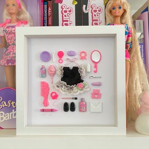 May include: A framed display of miniature doll accessories, featuring a black dress with lace, shoes, a comb, and a mirror. The collection includes pink and purple items, and is set against a backdrop of dolls and boxes on a shelf.