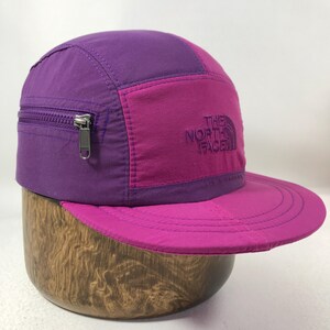 May include: A purple and magenta cap with a zippered pocket and a flat brim. The cap features the embroidered text "THE NORTH FACE" on the front. The cap is displayed on a wooden stand.