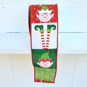 May include: A roll of wired ribbon featuring a repeating pattern of Christmas elf faces, striped elf legs, and candy canes. The design alternates between red and green backgrounds with white polka dots and festive holiday motifs throughout.