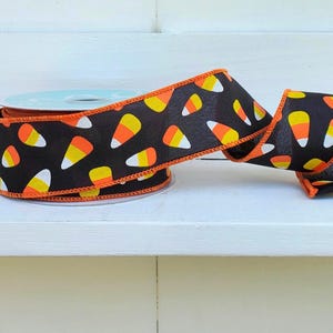 May include: A spool of black ribbon featuring a repeating pattern of candy corn shapes in yellow, orange, and white. The ribbon has a wired edge finished with bright orange stitching, providing structure to the fabric as it unrolls from the center.