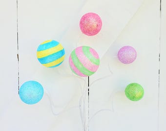 May include: A set of six cotton ball lights in various colors, including pink, blue, green, and yellow. The lights are strung together on a white wire and are hanging against a white wooden background.