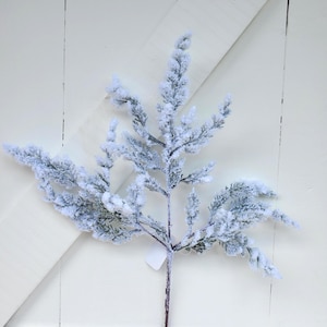 May include: A decorative artificial evergreen branch featuring a dense, flocked white coating that mimics the appearance of fresh snow. The branch has multiple smaller stems extending from a central stalk, with fine, needle-like foliage throughout.