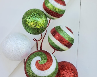 May include: A cluster of decorative Christmas ornaments in various colors. The ornaments are round and covered in glitter. Colors include red, green, white, and a swirl pattern. The ornaments are attached to a brown stem.