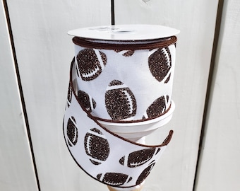 May include: A spool of white wired ribbon featuring a repeating pattern of brown glittery American footballs. The ribbon has dark brown stitched edges and is wound around a central plastic core, displaying a continuous print of sports balls.