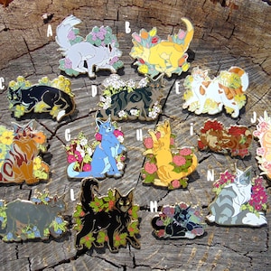 Op de afbeelding: Een set van 12 emaillepins met katten omringd door bloemen. De pins zijn in verschillende kleuren, waaronder blauw, groen, geel, oranje en wit. Elke pin heeft een ander kattenras en bloemendesign.