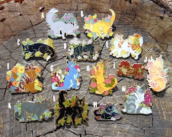 May include: A set of 12 enamel pins featuring cats surrounded by flowers. The pins are in various colors, including blue, green, yellow, orange, and white. Each pin features a different cat breed and floral design.