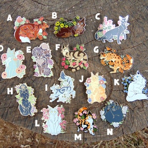 Puede incluir: Un conjunto de 14 pines de esmalte con diseños de gatos y flores. Los pines están dispuestos en una cuadrícula sobre una superficie de madera. Cada pin presenta una raza de gato y un diseño floral diferentes.