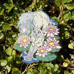 Puede incluir: Pin de esmalte detallado que representa un lobo estilizado descansando sobre nenúfares con flores rosas y centros amarillos. El lobo es de color gris claro con ojos azules, sobre un fondo azul y verde.