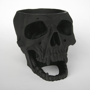 May include: A matte black skull-shaped container with a wide opening at the top and an open jaw. The container has detailed facial features, including eye sockets, a nose cavity, and teeth. The skull is a decorative item.