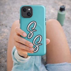 Puede incluir: Funda de teléfono turquesa con módulo de cámara negro y diseño de letra "S" en cursiva blanca y negra. La funda del teléfono tiene un agarre emergente incorporado. Una botella de agua verde es visible en el fondo.