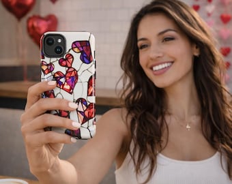 Puede incluir: Una funda blanca para smartphone con un diseño de corazón de vidriera en rojo, morado y rosa. La funda tiene un borde negro y la sostiene una persona. Globos rojos en forma de corazón en el fondo.