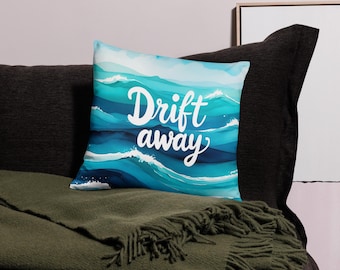 Puede incluir: Un cojín decorativo cuadrado con un diseño de acuarela de olas oceánicas en tonos azules y blancos. El cojín presenta las palabras "Drift away" en escritura blanca. El cojín está colocado sobre un sofá gris oscuro con una manta verde.