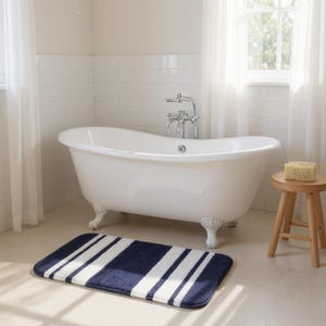 Op de afbeelding: Een witte badkuip met klauwpoten en een chromen kraan in een badkamer. Een marineblauwe en wit gestreepte badmat ligt op de vloer. Een kleine houten kruk heeft een spons.