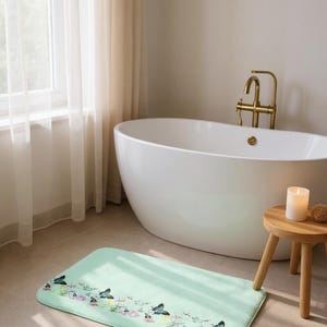 Könnte beinhalten: Eine rechteckige, mintgrüne Badematte mit einem Schmetterlings- und Blumenbordürendesign. Die Matte liegt vor einer weißen, ovalen Badewanne. Ein kleiner Holzschemel mit einer Kerze steht daneben.